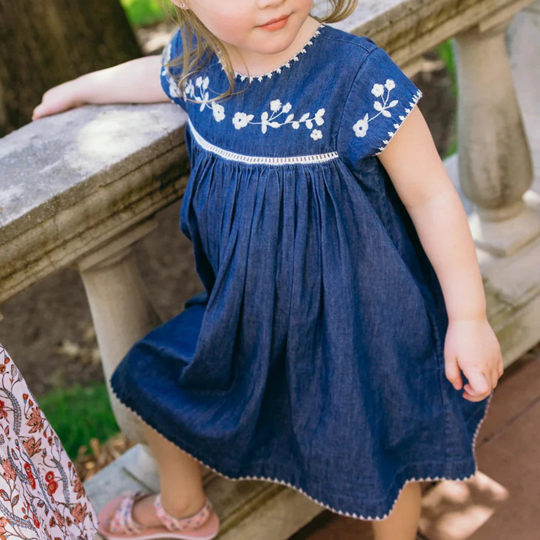 A young girl in the Minnow Girl's Chambray Embroidered Cap Sleeve Dress sits on a stone railing outdoors.