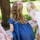Two young girls with long hair, one wearing the Minnow Girl's Chambray Embroidered Cap Sleeve Dress, lean over a stone railing outdoors.