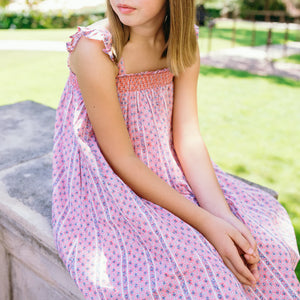 A young girl wears the Minnow Girls' Floral Tile Smocked Dress with Flutter Sleeves as she sits on a stone ledge outdoors, hands in her lap and face partially out of frame.