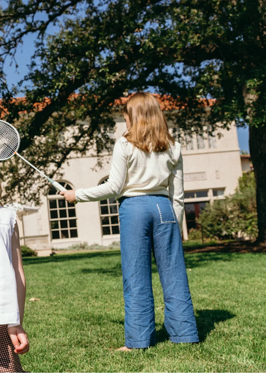 A girl wearing Minnow’s Girls' Chambray Embroidered Paperbag Pant stands on grass with a badminton racket, facing away from the camera; trees and a building appear in the background.