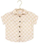 The Lou Lou & Company Baby Boys’ Cloud Muslin Shirt, featuring a beige and white checkered design with short sleeves and button-up front, hangs on a gold hanger against a white background—a versatile piece with a relaxed fit, ideal for casual days.