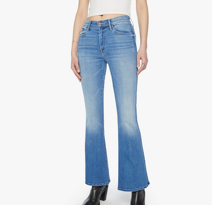 A person wears The Weekender flared denim jeans by Mother, paired with a white cropped top and black heeled boots, standing against a plain background.