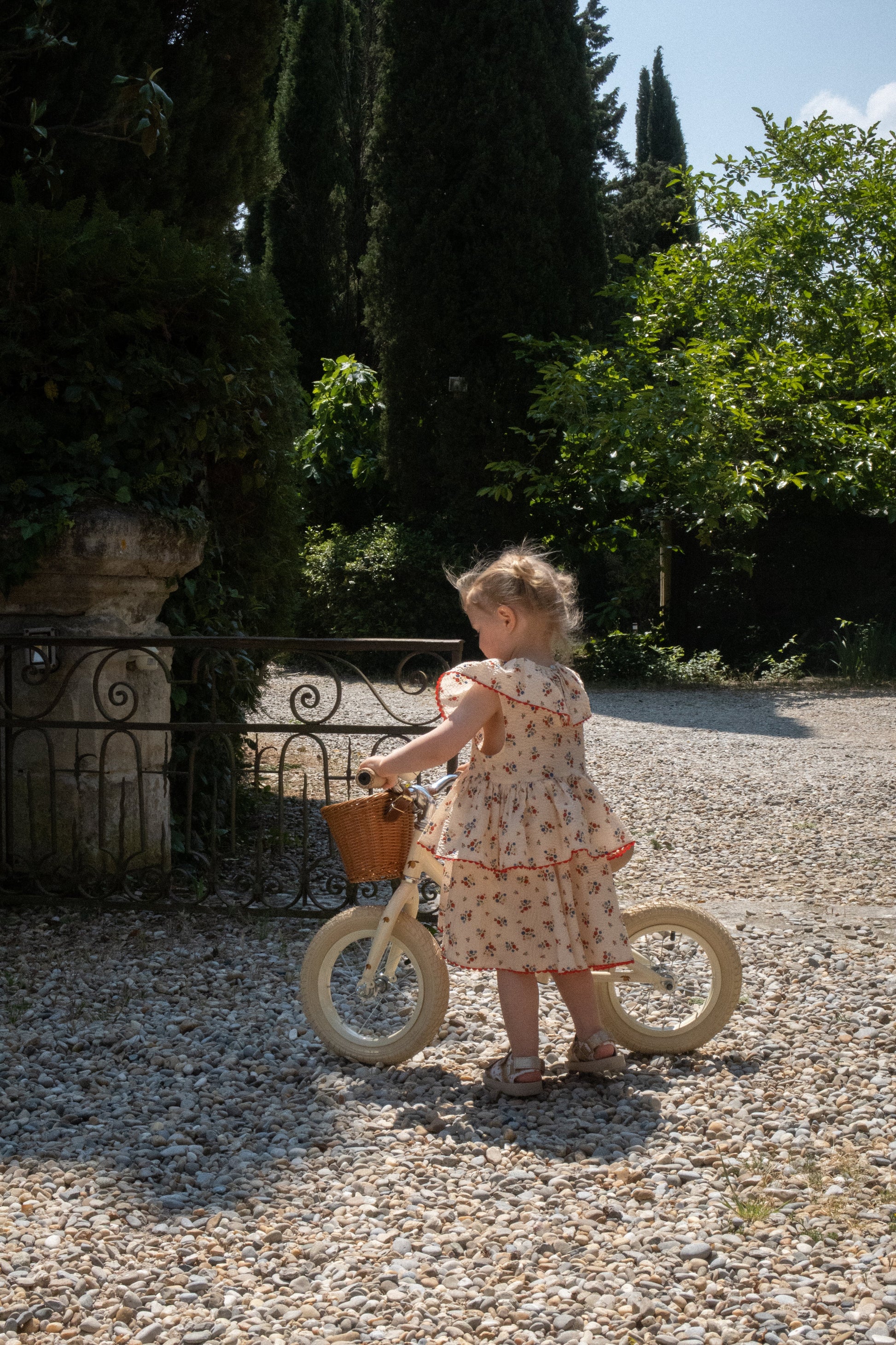 A young girl in a floral dress stands next to a Konges Slojd Balance Bike, featuring a sturdy steel frame, on a gravel path surrounded by greenery on a sunny day.