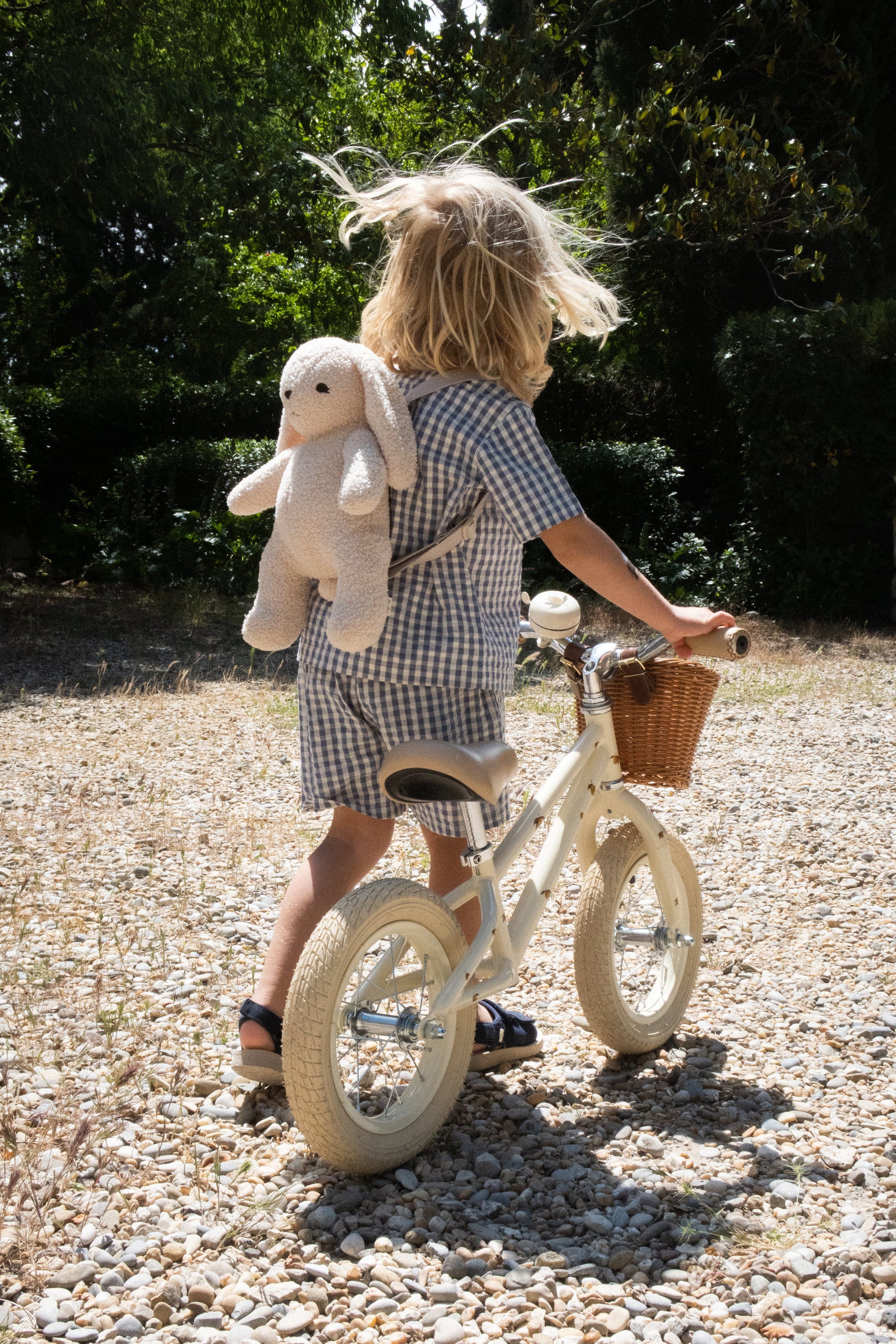 A child with long blonde hair rides a Konges Slojd Balance Bike on a gravel path. The lightweight design of the bike ensures easy handling, and the child’s blue and white checkered outfit complements their stuffed animal backpack perfectly.