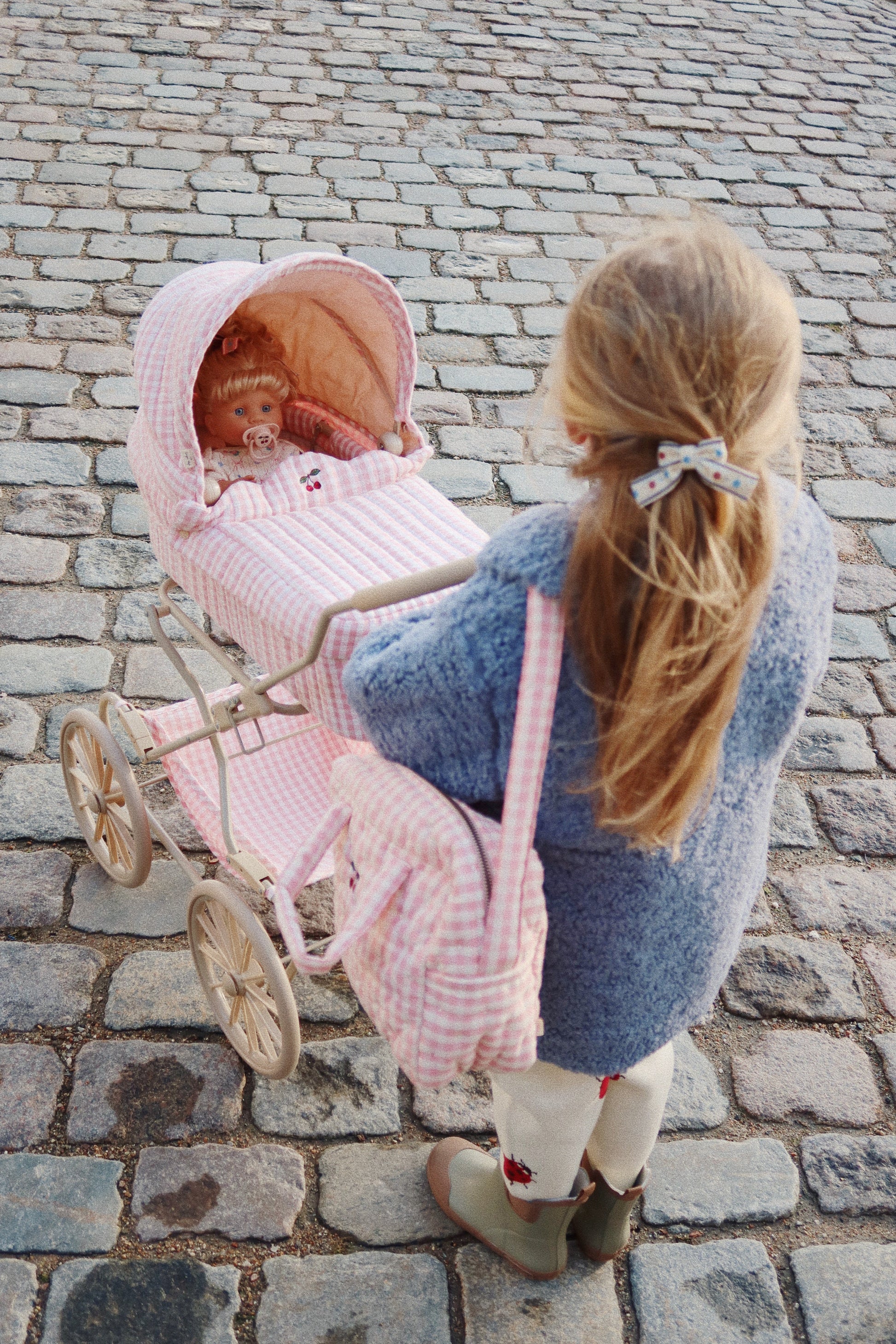 A young child with long blond hair, wearing a blue coat and white leggings, pushes a pink doll stroller with a baby doll inside on a cobblestone street. Over their shoulder hangs the Konges Slojd Doll Bag, made by the brand Konges Slojd, featuring an adjustable shoulder strap and crafted from 100% cotton.