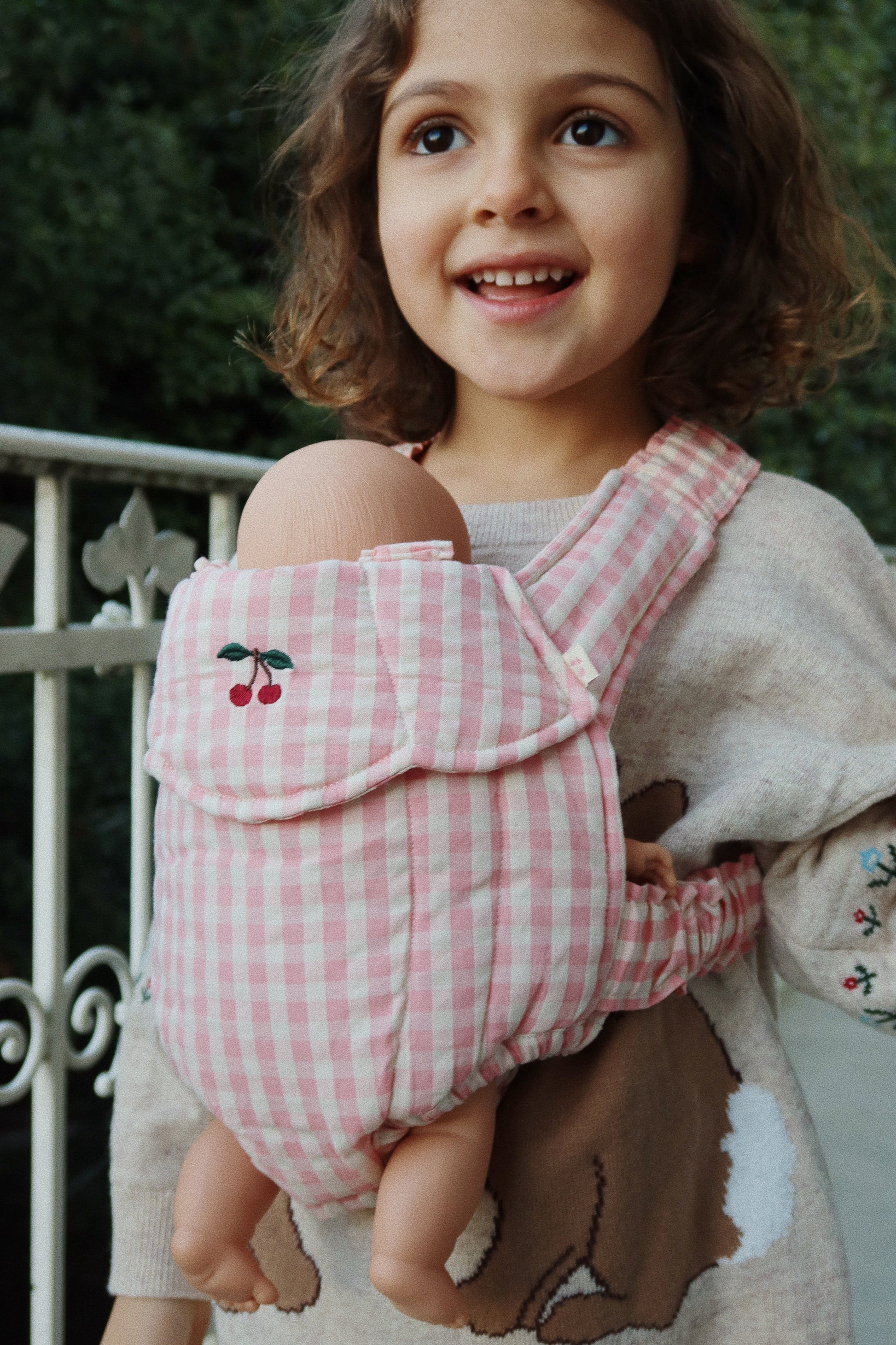 A child standing outdoors, smiling, while holding a doll in the Konges Slojd Doll Carrier—a pink and white checkered carrier made of 100% cotton—from the brand Konges Slojd on their chest.