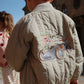 A child in a beige Konges Slojd Juno Bomber Jacket with a race car design stands outdoors. Another child is partly visible in the background, wearing a thermo jacket offering excellent insulation.