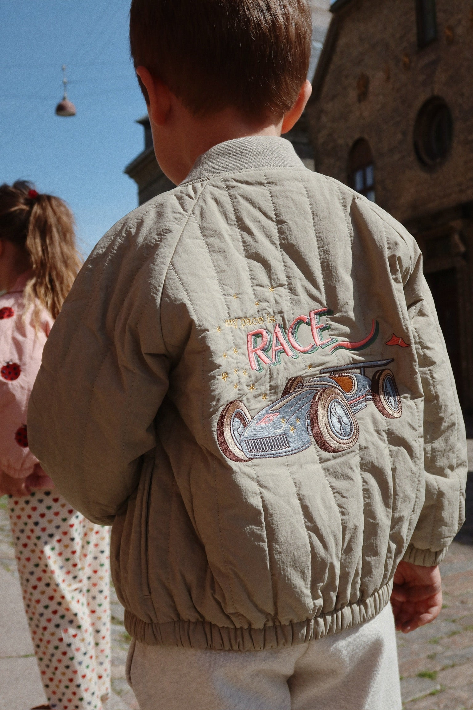 A child in a beige Konges Slojd Juno Bomber Jacket with a race car design stands outdoors. Another child is partly visible in the background, wearing a thermo jacket offering excellent insulation.