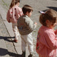 Three children in casual clothing walk down a cobblestone street. The child in the middle proudly showcases their Konges Slojd Juno Bomber Jacket, featuring a car design. This stylish piece captures attention and provides insulation, keeping them cozy and water-repellent as they explore.