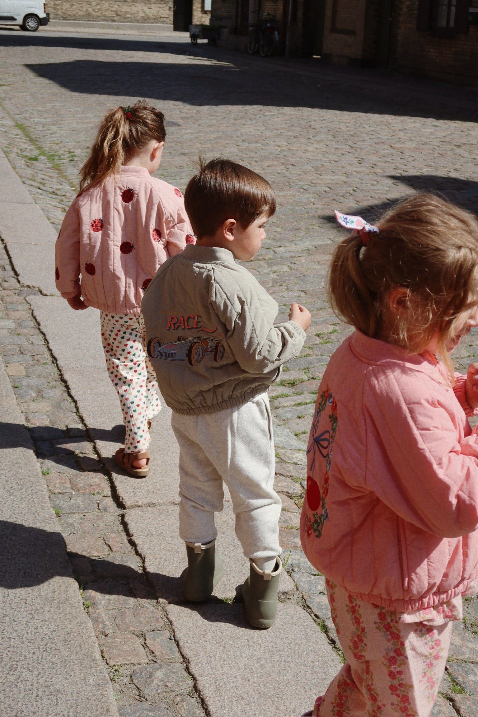 Three children in casual clothing walk down a cobblestone street. The child in the middle proudly showcases their Konges Slojd Juno Bomber Jacket, featuring a car design. This stylish piece captures attention and provides insulation, keeping them cozy and water-repellent as they explore.