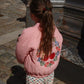A child with long hair stands facing away, wearing a pink Konges Slojd Juno Bomber Jacket adorned with floral and heart designs and heart-patterned pants, near a stone surface in the sun. The jacket’s water-repellent feature keeps them cozy and dry on any adventure.