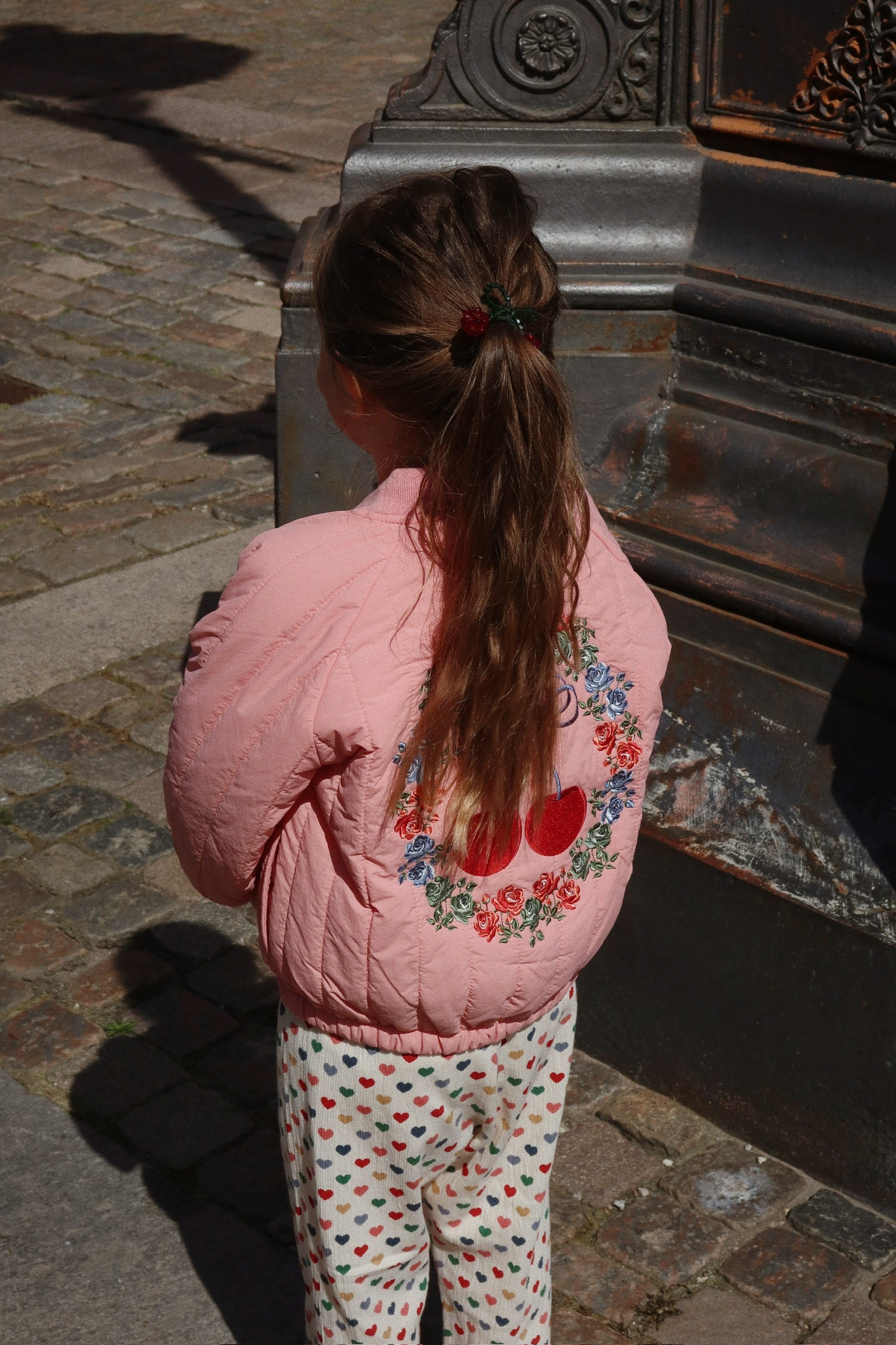 A child with long hair stands facing away, wearing a pink Konges Slojd Juno Bomber Jacket adorned with floral and heart designs and heart-patterned pants, near a stone surface in the sun. The jacket’s water-repellent feature keeps them cozy and dry on any adventure.