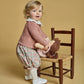 A blonde toddler in a 100% cotton pink Kidiwi Baby Girls' Myrtille Smocked Bloomer stands by a wooden chair, holding a pink-dressed doll. She wears floral shorts, white tights, and white shoes.