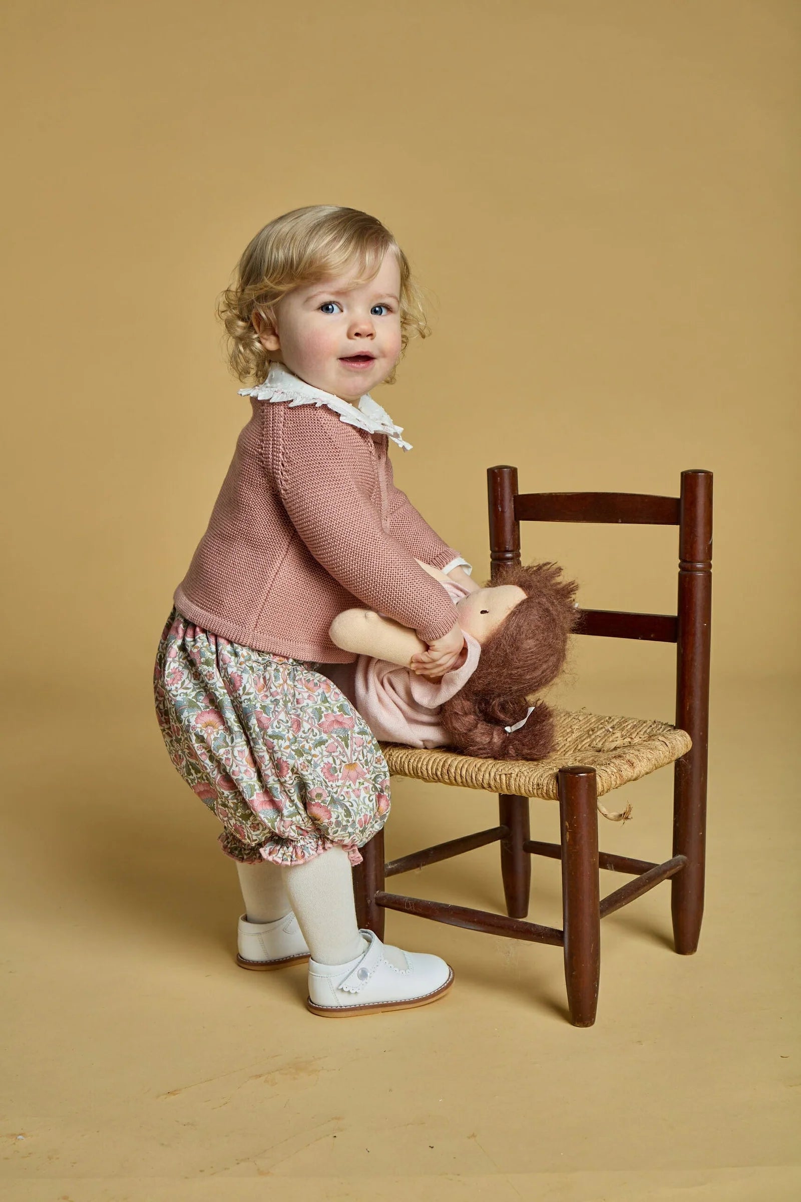 A blonde toddler in a 100% cotton pink Kidiwi Baby Girls' Myrtille Smocked Bloomer stands by a wooden chair, holding a pink-dressed doll. She wears floral shorts, white tights, and white shoes.