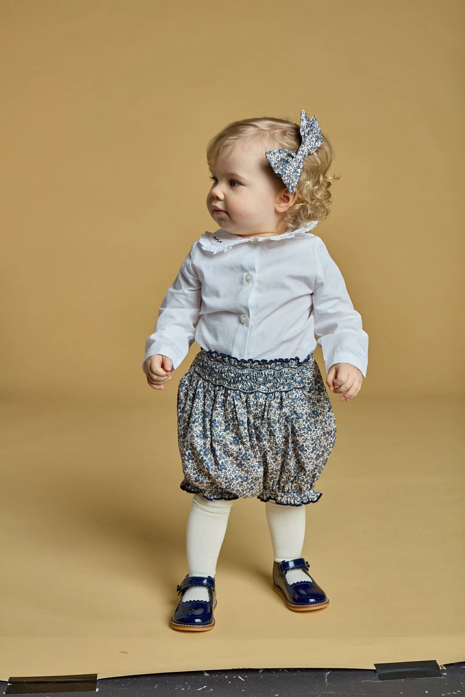 A young child stands against a beige background, dressed in a white shirt, Kidiwi Baby Girls' Myrtille Smocked Bloomer with a navy blue print, white tights, blue shoes, and a matching floral hair bow.