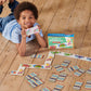 A child smiles on a wooden floor, holding up a domino card while playing Eeboo's Dinosaur Head to Toe Dominoes with colorful cards spread out beside the game box.