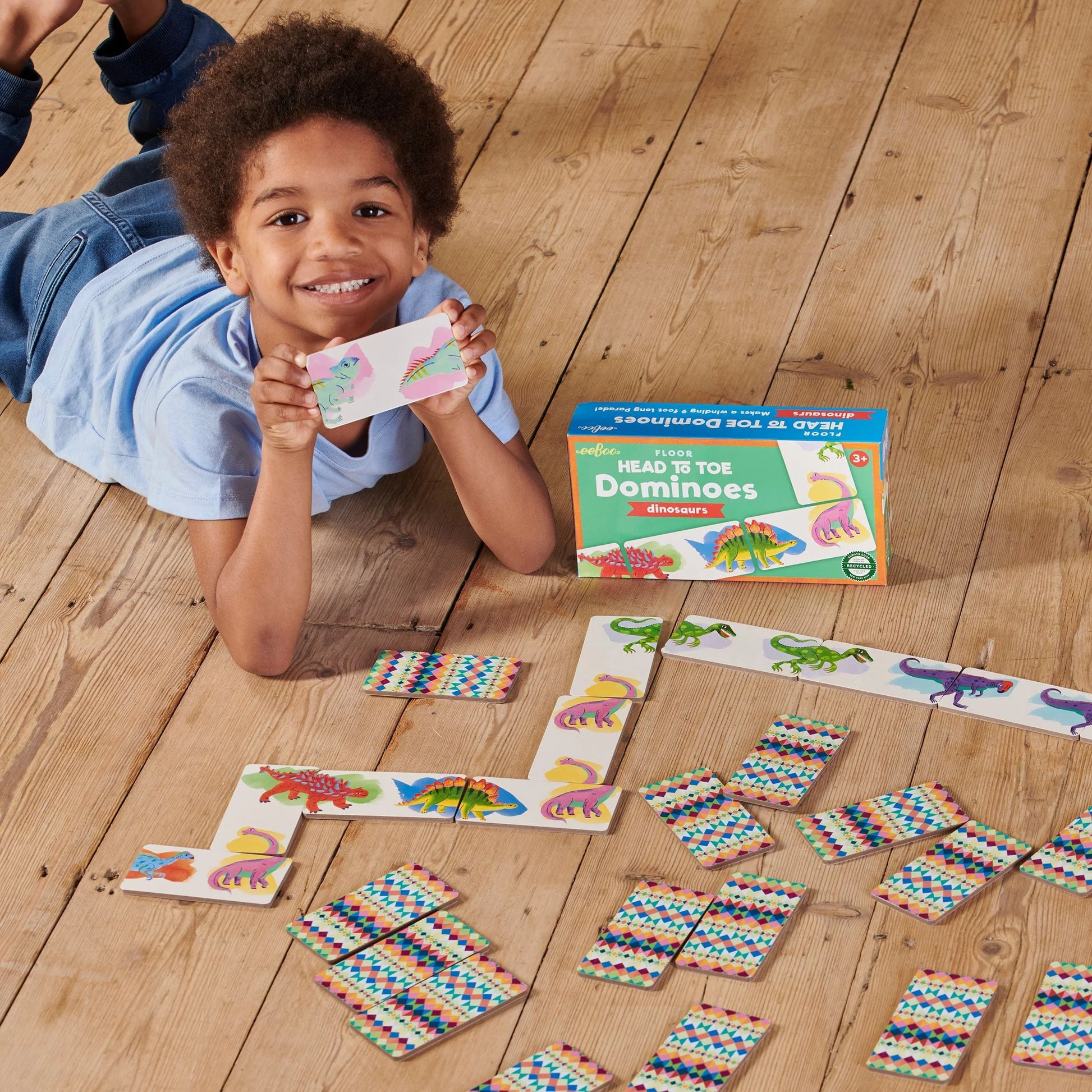 A child smiles on a wooden floor, holding up a domino card while playing Eeboo's Dinosaur Head to Toe Dominoes with colorful cards spread out beside the game box.