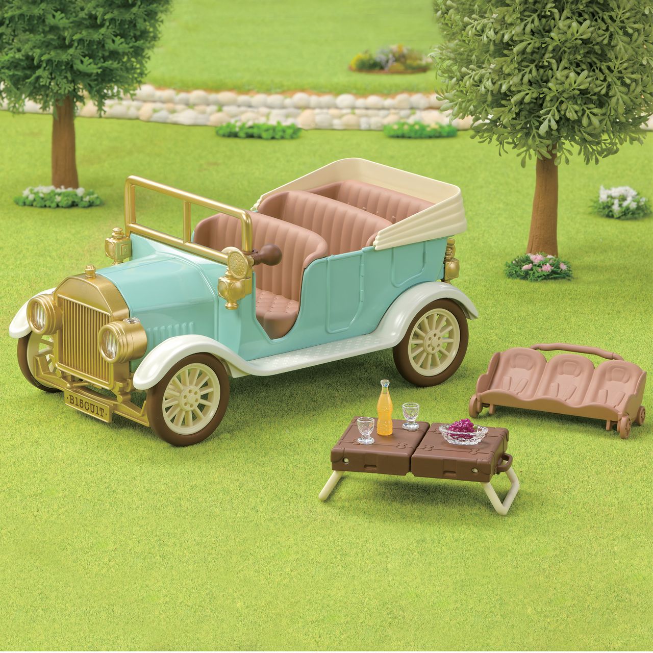 A Calico Critters Classic Family Car is parked on the grass beside a folded seat that fits 10 figures, next to a picnic table with drinks and a tray of food. Trees and a stone path appear in the background.