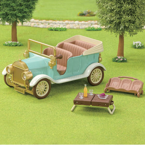 A Calico Critters Classic Family Car is parked on the grass beside a folded seat that fits 10 figures, next to a picnic table with drinks and a tray of food. Trees and a stone path appear in the background.