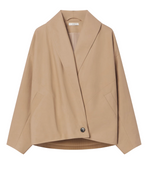 The CO Wrap Jacket is a beige, long-sleeve, collarless design with a cocooned silhouette and a large shawl lapel. It features an asymmetric button closure and is displayed on a hanger against a white background.
