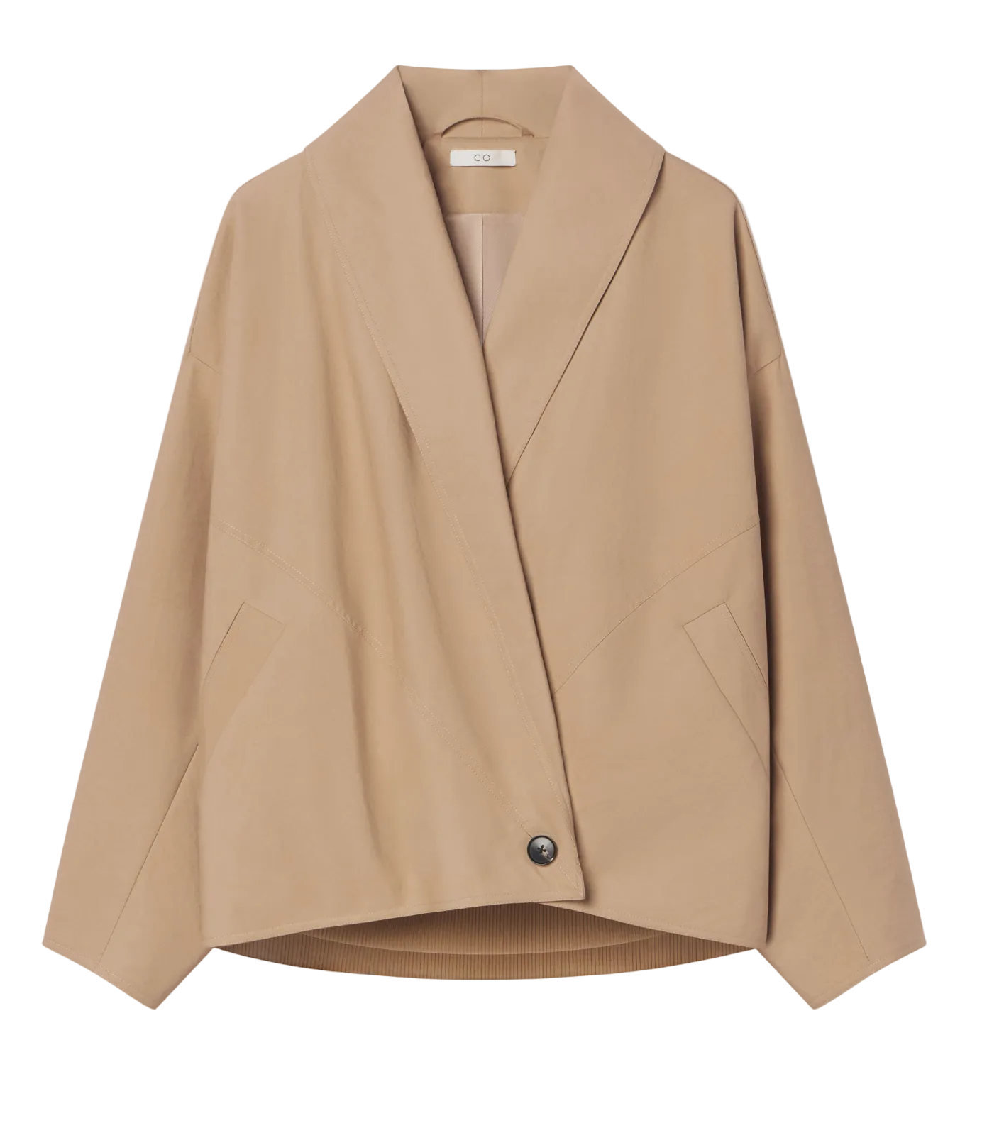 The CO Wrap Jacket is a beige, long-sleeve, collarless design with a cocooned silhouette and a large shawl lapel. It features an asymmetric button closure and is displayed on a hanger against a white background.