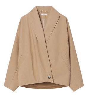 The CO Wrap Jacket is a beige, long-sleeve, collarless design with a cocooned silhouette and a large shawl lapel. It features an asymmetric button closure and is displayed on a hanger against a white background.