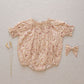 The Noralee Baby Girls' Maddie Romper features short sleeves and delicate floral embroidery, styled with a matching bow and two dried grasses, all arranged on a neutral fabric background.