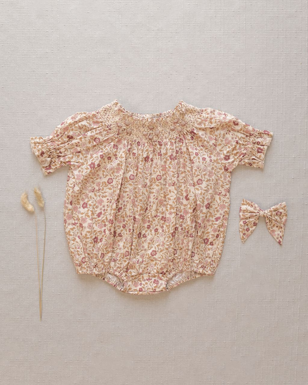 The Noralee Baby Girls' Maddie Romper features short sleeves and delicate floral embroidery, styled with a matching bow and two dried grasses, all arranged on a neutral fabric background.