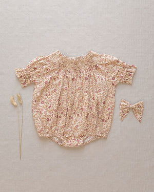 The Noralee Baby Girls' Maddie Romper features short sleeves and delicate floral embroidery, styled with a matching bow and two dried grasses, all arranged on a neutral fabric background.