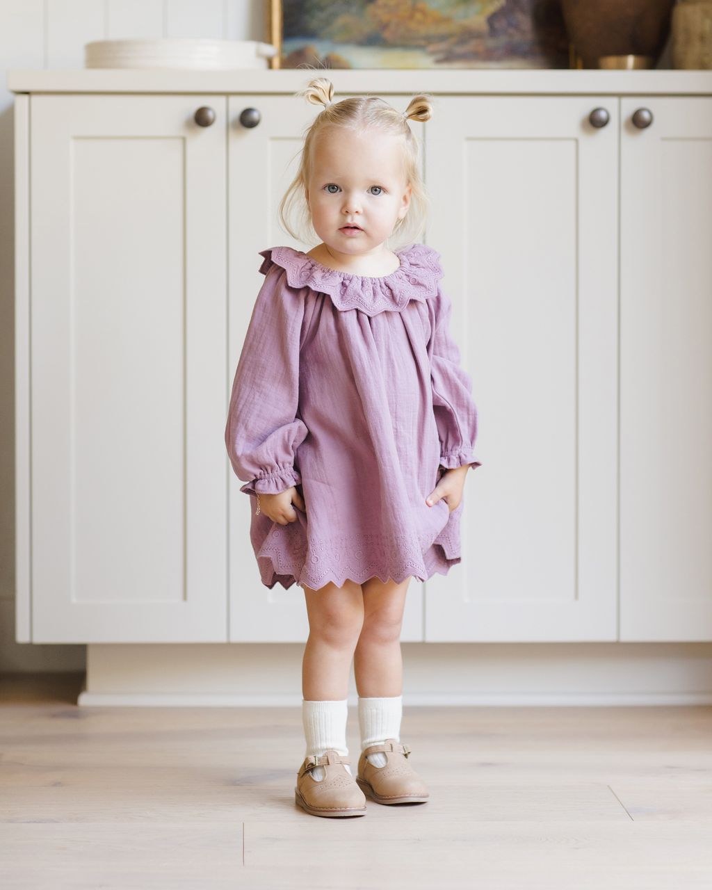 A young blond girl with pigtails stands indoors in front of a white cabinet, wearing the Quincy Mae Baby Girls' and Girls' Long Sleeve Isla Dress with white socks and tan shoes.