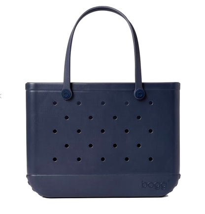 The Original Bogg® Bag by Bogg Bags is a navy blue tote featuring perforated holes, two sturdy handles, and a rubber-like construction. This stylish, washable beach bag has “Bogg” embossed on the lower right corner.