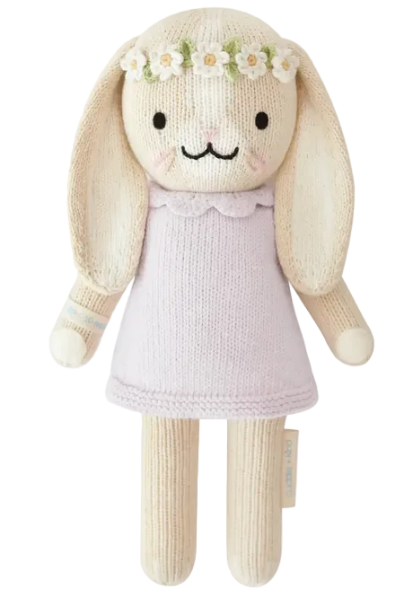 Hannah the Bunny Violet - Little 13" by Cuddle + Kind features a knitted bunny doll with long ears, dressed in a light purple dress and adorned with a white flower crown. Standing upright against a plain background, this charming plush toy makes an inspirational gift for any special occasion.