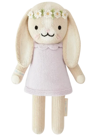 Hannah the Bunny Violet - Little 13" by Cuddle + Kind features a knitted bunny doll with long ears, dressed in a light purple dress and adorned with a white flower crown. Standing upright against a plain background, this charming plush toy makes an inspirational gift for any special occasion.