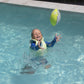 A child wearing a life jacket stands in a pool, reaching up with both hands to catch the Sunnylife Tides Out Citrus Neoprene Football in mid-air.