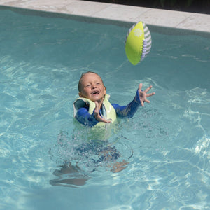 A child wearing a life jacket stands in a pool, reaching up with both hands to catch the Sunnylife Tides Out Citrus Neoprene Football in mid-air.