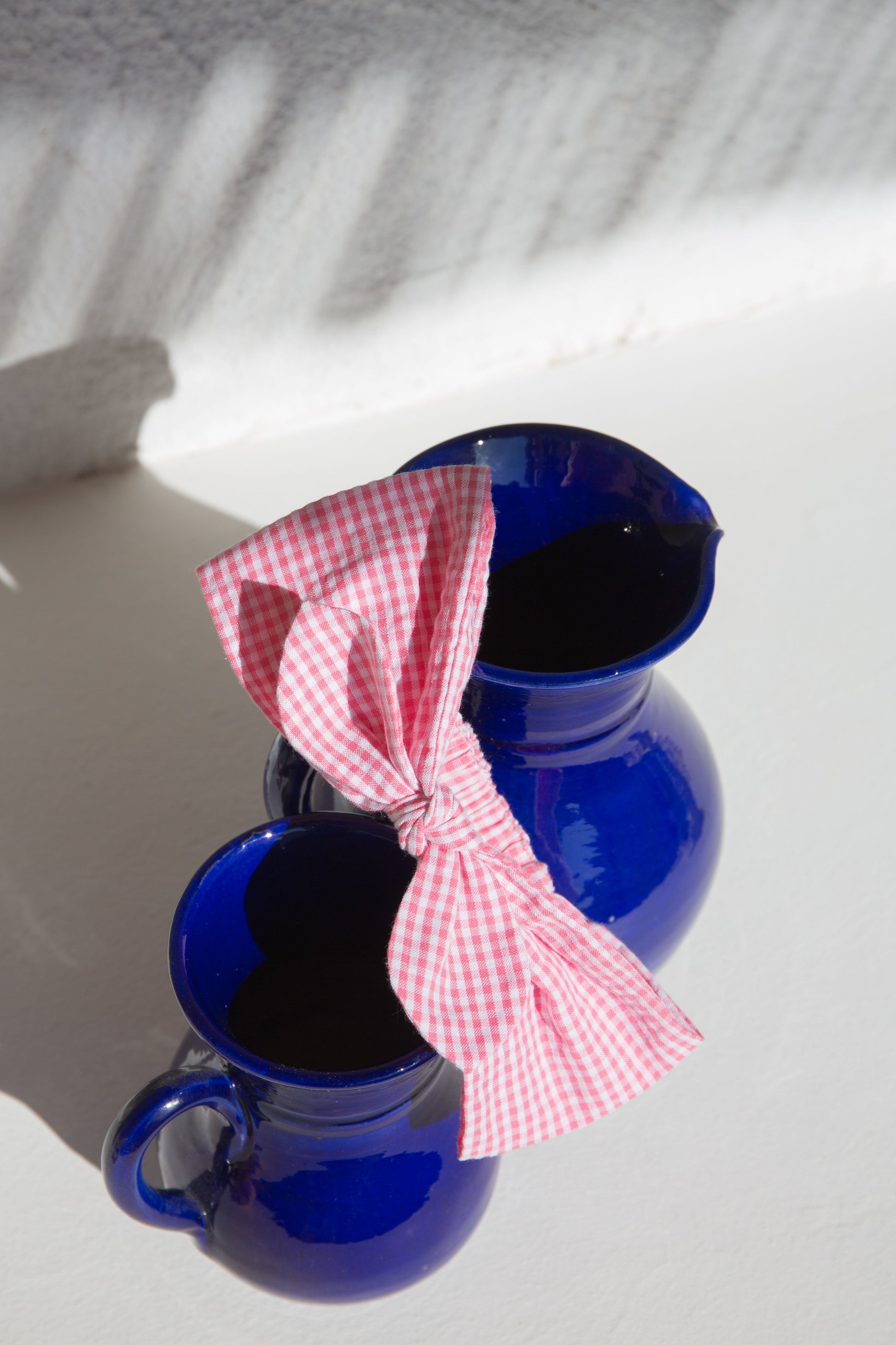 Two blue ceramic pitchers, one with a Louise Misha Baby Cally Headband in a pink fragola bow design, sit on a sunlit white surface. Shadows form patterns around these charming decor pieces perfect for delighting little girls.