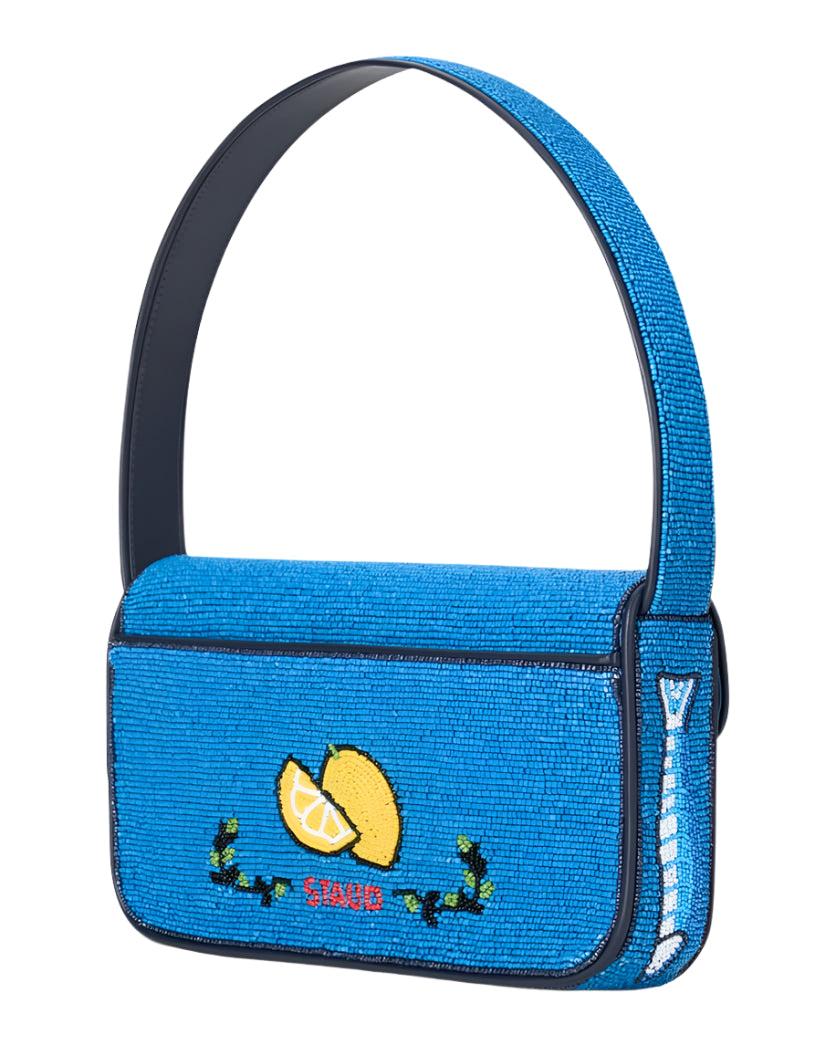 The Staud Tommy Beaded Bag in blue features a "SAVOR" lemon slice design, crafted from vegan leather with hand-beaded floral accents and a thick matching strap—making it stand out beautifully against any background.