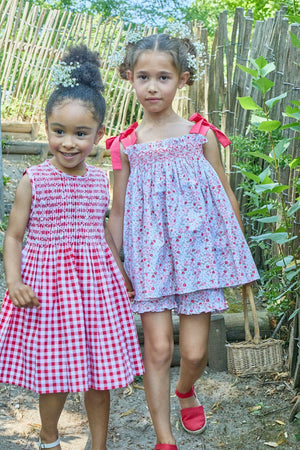 Two young girls stand outdoors by a wooden fence. One wears a red checkered dress, the other a floral dress with red bows and Kidiwi's Baby Girls' and Girls' Nolana Short in Cherry Red Print. Both have flowers in their hair.