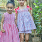 Two girls stand by a wooden fence; one wears a cherry red print dress, while the other wears the Kidiwi Baby Girls' and Girls' Sariette Smocked Blouse in floral 100% cotton with red ribbons. Both have flowers in their hair.