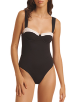 A woman wearing the Evarae Nova One Piece, a black swimsuit with white trim featuring supportive underwire and adjustable straps, stands with her arms relaxed at her sides.