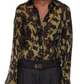 A person wears the Victoria Beckham Pyjama Shirt, styled as a sheer olive green and black floral blouse, paired with dark high-waisted pants and a rectangular belt buckle.