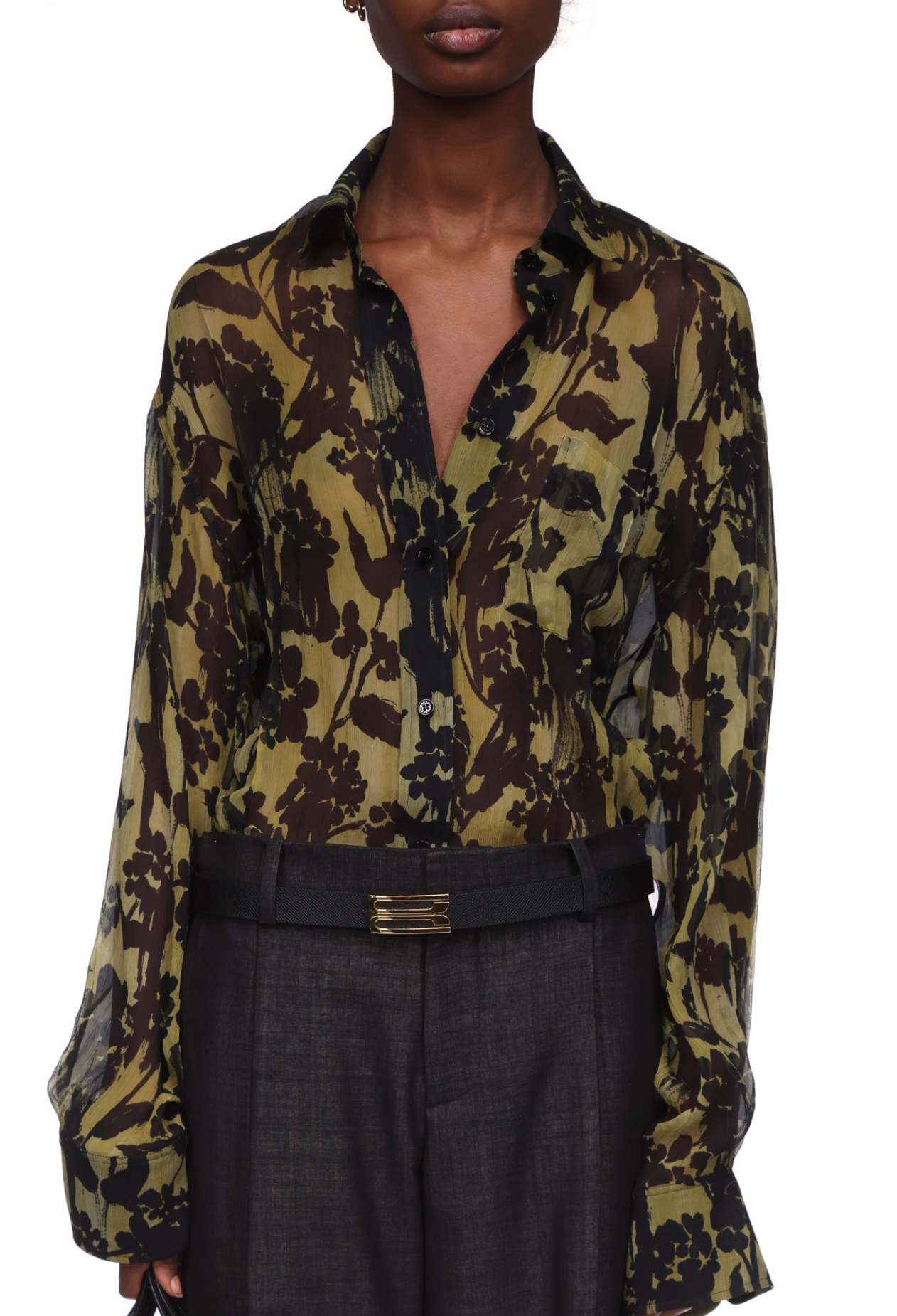 A person wears the Victoria Beckham Pyjama Shirt, styled as a sheer olive green and black floral blouse, paired with dark high-waisted pants and a rectangular belt buckle.
