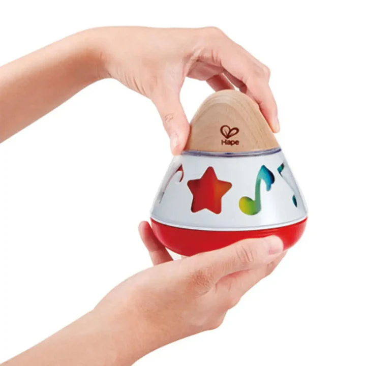 A person holds a Hape Rotating Music Box, a baby toy featuring a wooden top, silver middle decorated with colorful shapes, and a red base that plays soothing music.