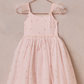 The Girls' Poppy Dress by Noralee is a delightful pink sleeveless creation with feathered shoulder straps and a charming polka dot pattern, elegantly displayed on a copper hanger against a beige backdrop.