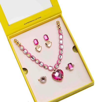 A yellow box from Super Smalls displays the Perfectly Pink Mega Set, featuring play jewelry including a gemstone heart pendant necklace, pink stud earrings, gold heart earrings, a pink gem ring, and a round pink gem brooch inside.