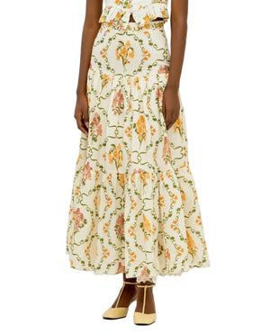 A person wears the Anis Skirt by Agua By Agua Bendita, a long cream-colored tiered skirt with a floral pattern and lace hem, paired with light yellow shoes.