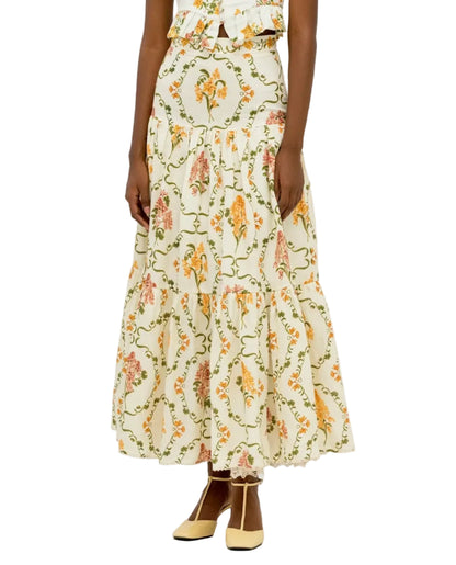 A person wears the Anis Skirt by Agua By Agua Bendita, a long cream-colored tiered skirt with a floral pattern and lace hem, paired with light yellow shoes.