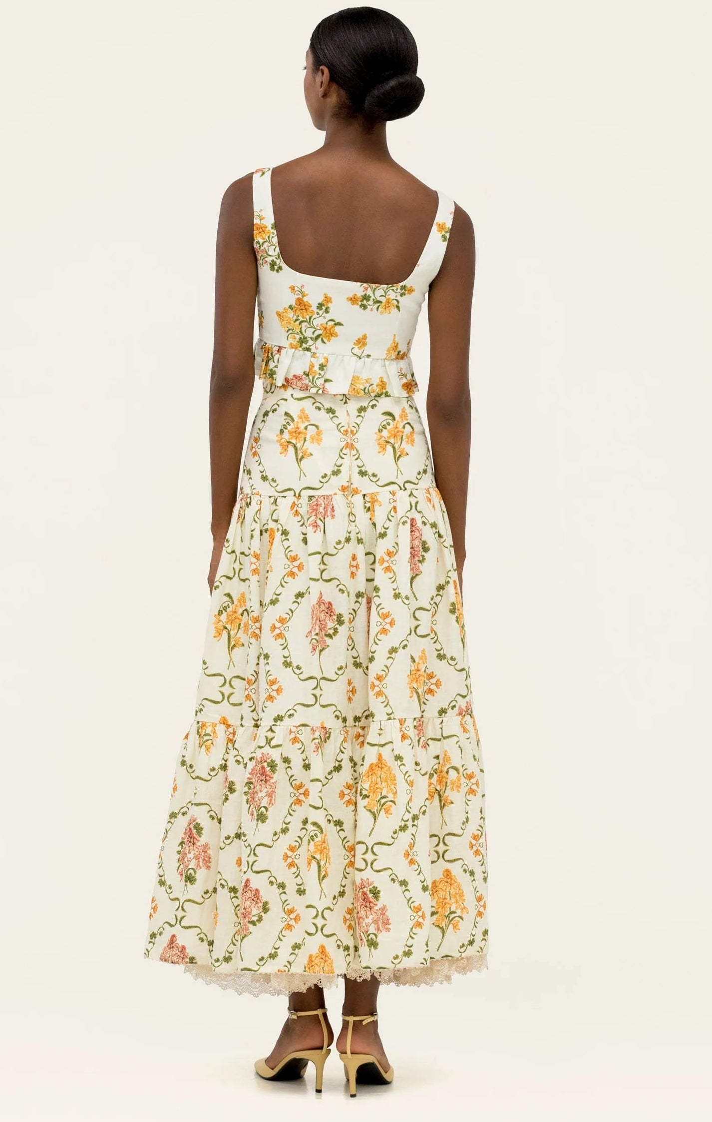 A woman faces away, wearing the Anis Skirt by Agua By Agua Bendita—a white maxi skirt with colorful floral patterns and a lace hem. She pairs it with gold heels and styles her hair in a neat low bun.