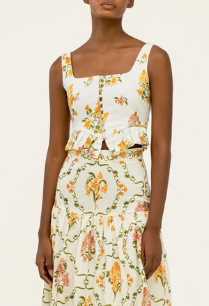 A woman wears the Anis Skirt by Agua By Agua Bendita, a sleeveless, square-neck top and slim-fit linen skirt set in white with yellow and pink floral patterns.