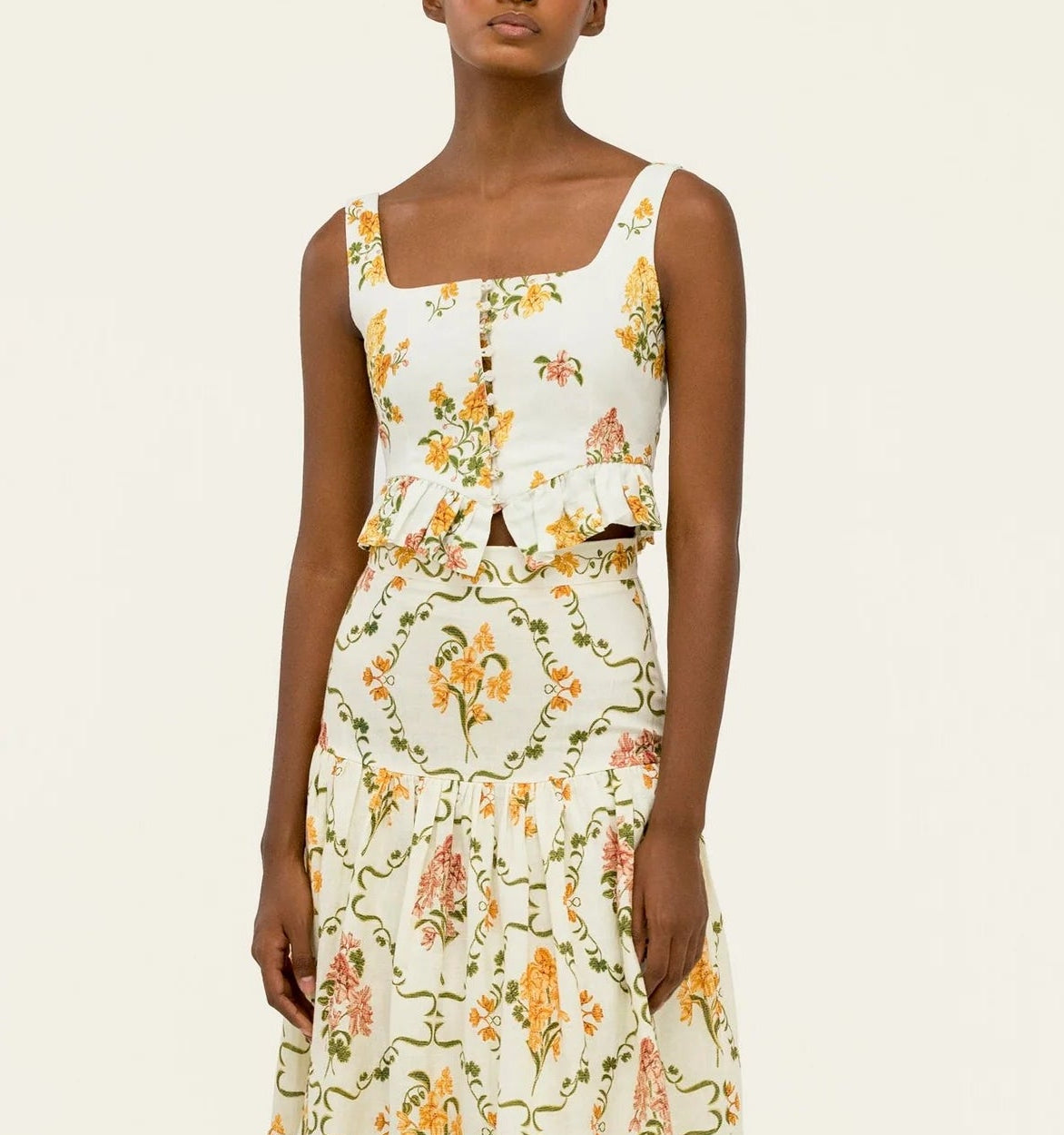 A woman wears the Eslabon Top by Agua By Agua Bendita—a sleeveless, square-neck top with ruffle and button details—paired with a matching floral skirt in yellow, orange, and green tones.